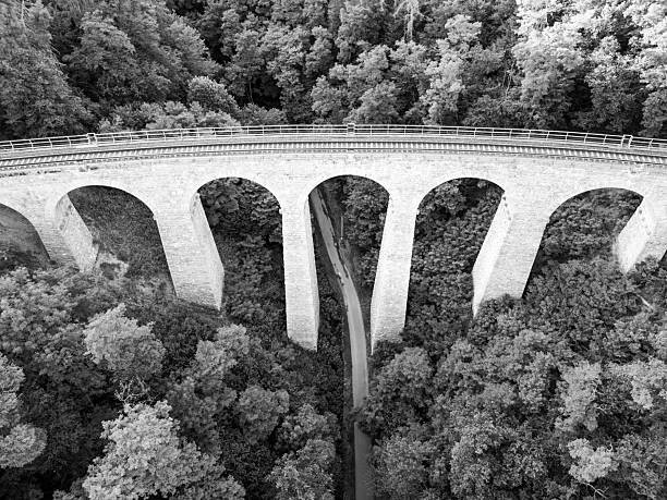 The historic Zampach stone railway bridge arches gracefully over a lush forest, showcasing its impressive stonework and connecting the rail tracks to the verdant landscape below.