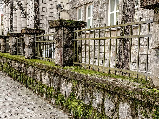 Moss Covered Stone Fence Along Belgrade Sidewalk, Weathered Sandstone Blocks Topped With Iron Railing, Thick Green Moss In Joints, Damp Pavement, Bare