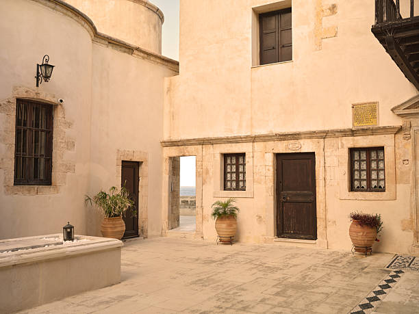 Greek Catholic Monastery: Photo from the island of Crete (Greece): A sunlit courtyard with beige stone walls, dark wooden doors, and potted plants. An arch leads to the sea, while windows and a balcony add character. The scene conveys warm Mediterranean charm, history, and tranquil everyday life in Greece.