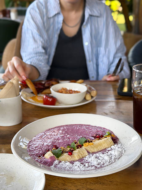 Close-up image of fresh berry smoothie bowl for healthy breakfast with blueberries, banana, pomegranate, granola, oats, coconut, unrecognisable woman eating fried breakfast al fresco dining setting