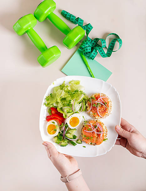 Breakfast with 2 sandwiches with salmon, boiled egg and fresh vegetable salad. The plate is held by female hands, on the table there is a notebook, pen, dumbbells and measuring tape. Flat lay