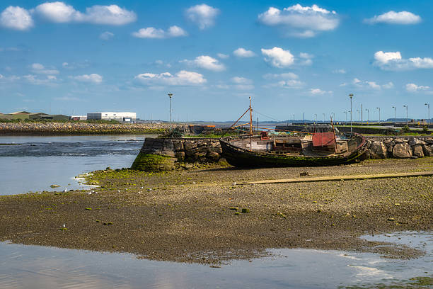 Low tide harbor stranded wooden trawler grounded on rocky mudflat, distant breakwater and calm sky, weathered paint, seaweed textures, quiet coastal solitude and nostalgic atmosphere