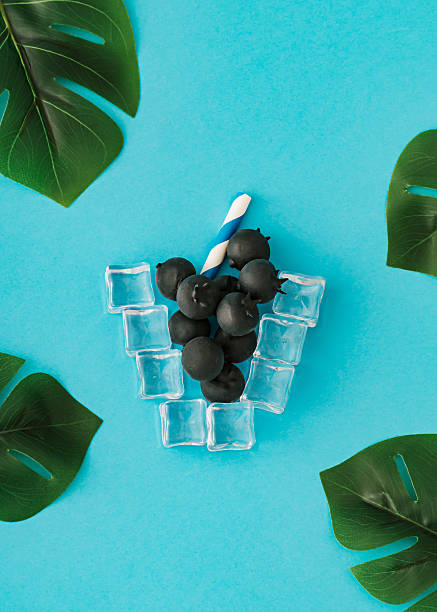 Fresh blueberries with ice cubes and paper straw on blue background with tropical leaves. Summer drink concept.