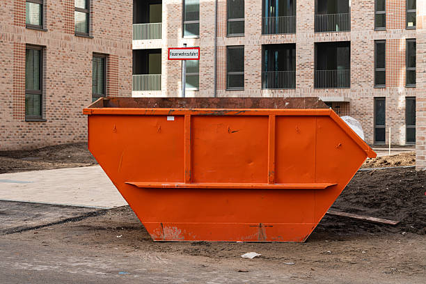 A large orange dumpster sits at a construction site next to new residential buildings. It is empty and surrounded by dirt and materials, typical of ongoing construction work.