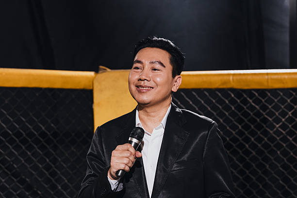 Man speaking with microphone at an event in a venue with a cage in the background while people watch.