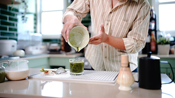 young asian man preparing iced matcha latte pouring green matcha tea into a glass with milk, creating a refreshing iced matcha latte in a cozy kitchen setting