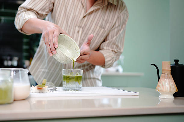 young asian man preparing iced matcha latte pouring green matcha tea into a glass with milk, creating a refreshing iced matcha latte in a cozy kitchen setting