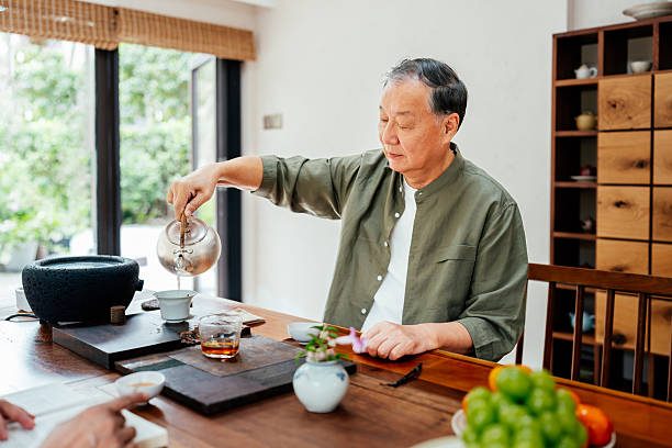 Asian senior man pouring traditional Chinese tea in a cup and enjoying afternoon tea at home. Traditional Chinese culture. Concept of hygge and slow life for senior people.