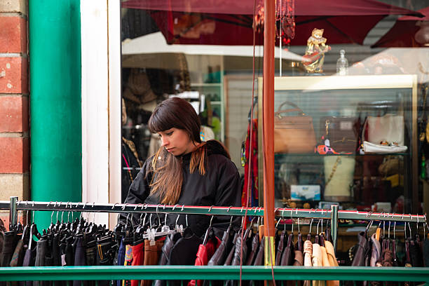 Young woman browsing clothing on a rack at a flea market in Paris, France. The candid lifestyle scene shows a shopper carefully examining garments at a second-hand clothing stall inside an urban market. The image highlights vintage fashion culture, sustainable shopping, and the everyday atmosphere of Paris flea markets where visitors explore unique clothing and retro styles.