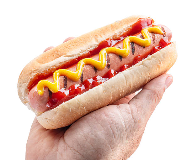 Classic American hot dog with a grilled sausage, ketchup and yellow mustard, served in a soft bun in a man's hand isolated on a white background.