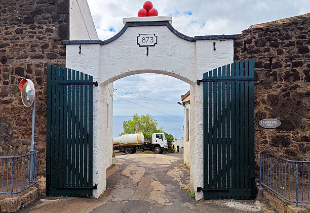 Half Tree Hollow, Saint Helena: main entrance gate to Ladder Hill Fort overlooking Jamestow. A white-painted masonry archway dating to 1873, topped with a decorative arrangement of three red cannon balls. The arch is flanked by substantial defensive walls constructed from rough, dark volcanic stone, reflecting the site's original military function. Dark green wooden slatted gates stand open, revealing an asphalt path that leads toward a clear view of the South Atlantic Ocean on the horizon. Historically, this fortification was developed by the British East India Company and subsequently reinforced by the British military to defend the anchorage below.