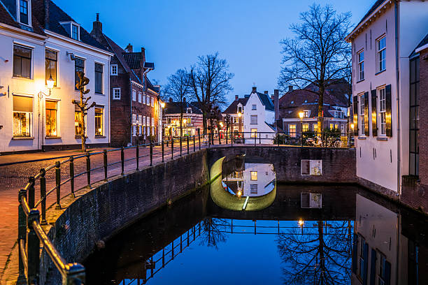 Amersfoort, The Netherlands – February 19, 2026: Atmospheric evening scene of a small brick bridge over a canal in the historic city center of Amersfoort, with traditional Dutch houses, warm street lighting and reflections in the calm water.