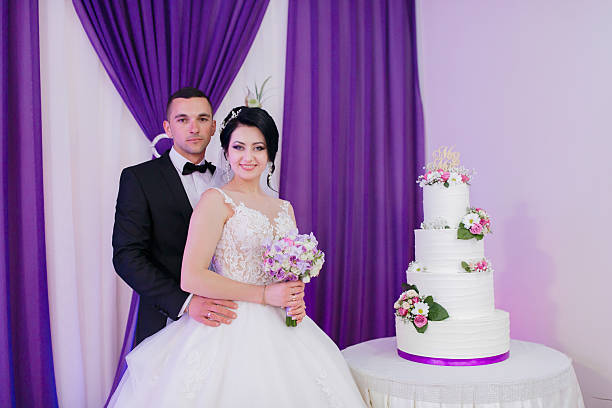 A bride and groom pose in front of a wedding cake