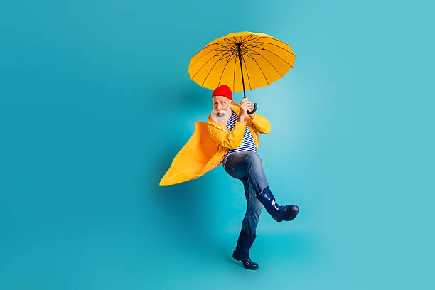 A cheerful senior man wears a bright yellow raincoat striped shirt jeans and boots while he dances with a yellow umbrella against a vivid blue background creating a fun and dynamic rainy day mood