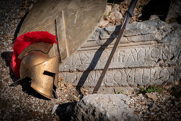 Ancient Greek Spartan helmet with red plume, sword and shield resting against stone ruins outdoors