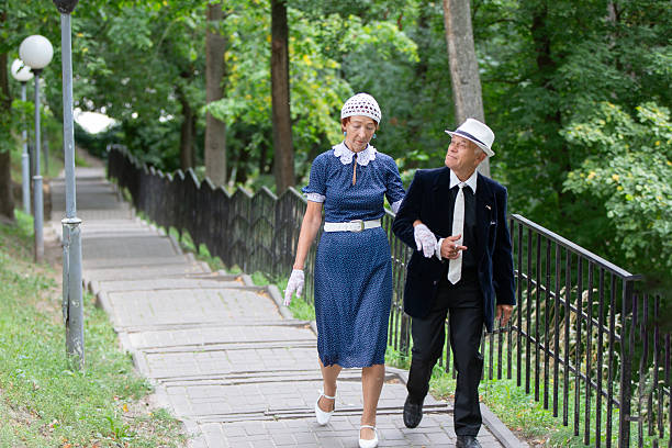 An elderly couple strolls closely together along a tree-lined pathway. They wear stylish attire, enjoying the warm sunlight and vibrant surroundings, reflecting joy and companionship.