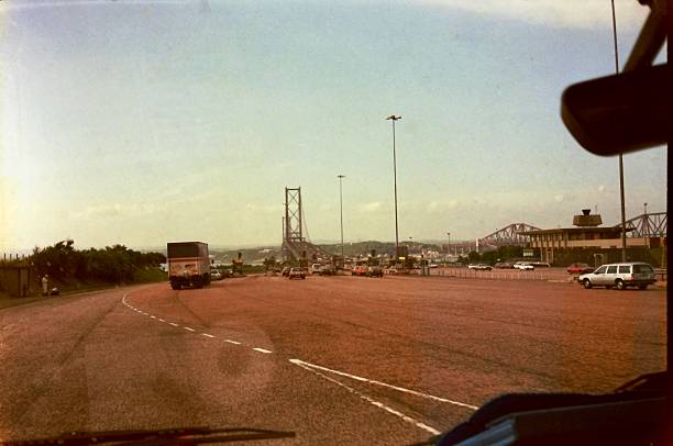 Forth Road Bridge 1984, shot from a van window, approaching toll-booths with distinctive red road tarmac. To the right is the  Forth Bridge railway crossing. Fife county, Scotland. Headed north, from Edinburgh to Fife, crossing the Firth of Forth. restored from an original 35mm film negative. Just visible in the van window is the reflection of a toilet roll on the dashboard. The van's rear view mirror is top right.