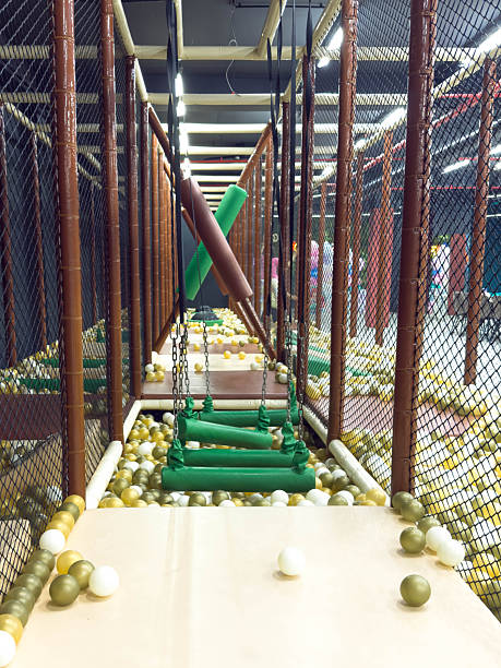 Indoor children adventure park with suspended green soft obstacles and chain steps above colorful ball pit, safe play zone surrounded by protective netting, active entertainment and family leisure concept