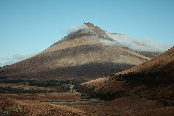 Mountain peak rising above a valley in the Scottish Highlands with clouds drifting over the summit. Scenic natural landscape with rugged terrain and soft light.