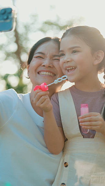 Joyful girl making soap bubbles while her mother takes pictures in the park