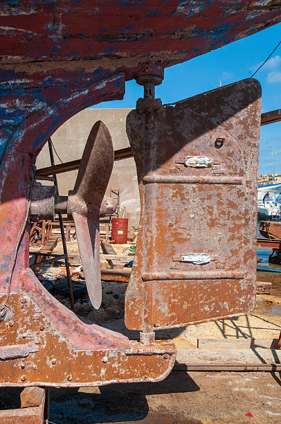 Close-up shot of a boat's rusty rudder and propeller, dry-docked for maintenance. The texture of corroded metal and peeling paint tells of years at sea, captured within the industrial atmosphere of a busy shipyard under a clear sky.