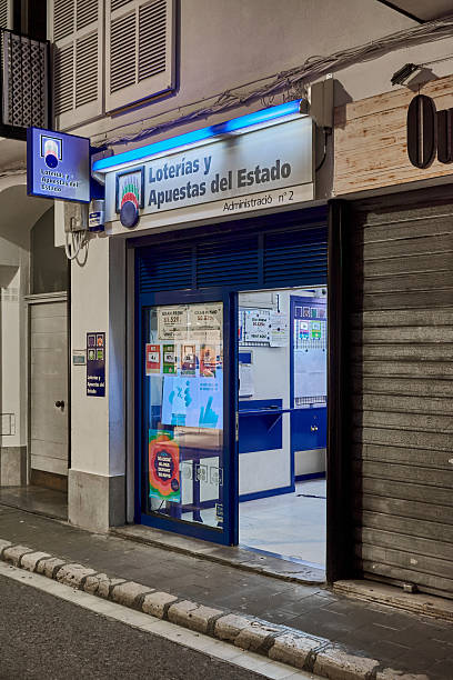 Sitges. Spain - March 13, 2026: Exterior view of a Loterias y Apuestas del Estado office at night with illuminated blue signage and lottery advertisements on the glass door.