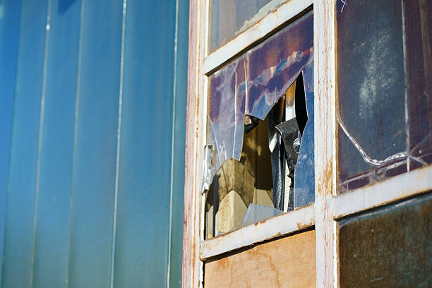 Old window with shattered plastic panel and peeling paint on facade. Neglect, building damage, repair concept and industrial urban background.