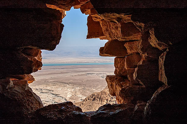 Israel, Masada : Panoramic view from inside the ancient Masada fortress in the Judean Desert, through a breach in the rock wall revealing the vast desert landscape and the turquoise waters of the Dead Sea below.