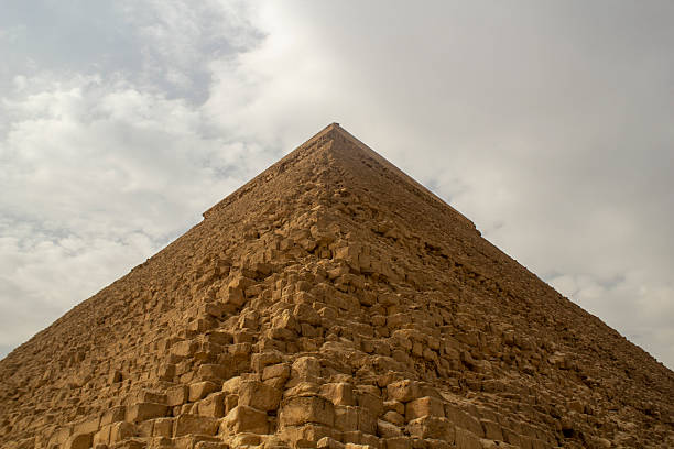 A striking low-angle perspective looking up at the monumental Pyramid of Khafre (Chephren) in Giza, Egypt. The image vividly captures the massive, weathered limestone blocks leading up to the original smooth casing stones preserved at the apex. Set against a dramatic overcast sky, this composition emphasizes the awe-inspiring scale, texture, and architectural majesty of this ancient wonder. Ideal for historical, archaeological, and travel-related visual projects.