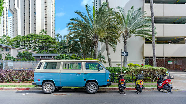 Waikiki, Honolulu, Hawaii, March 7, 2026:  A vibrant blue-and-white classic Volkswagon bus, likely a 1970's bay window camper (T2model), parks curbside in sunny Waikiki, Honolulu, Hawaii, surrounded by swaying palm trees, high-rise condos, colorful scooters, and lush greenery - evoking a pure island wanderlust.