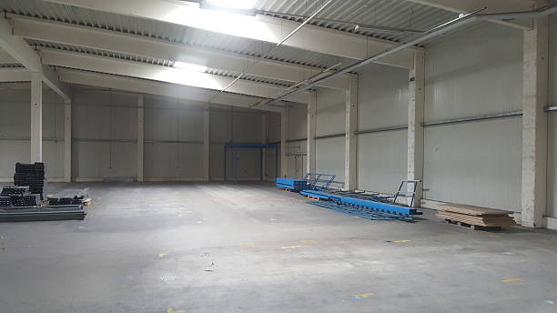 An empty, large industrial warehouse space with high ceilings, skylights, and some construction materials on the floor awaiting assembly or storage