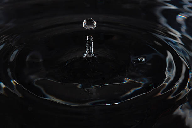 179.Water droplet mid-air above surface with splash column and concentric ripples. Low lighting and reflections highlight fluid motion and surface tension.