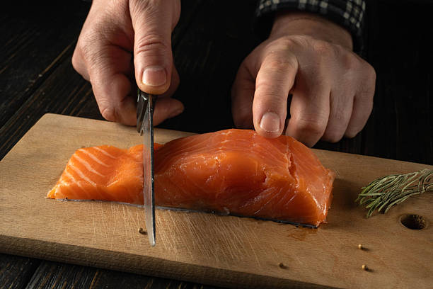 A person is using scissors to cut a salmon fillet on a wooden board. The kitchen has a rustic look with dark wood. Fresh herbs are nearby for cooking