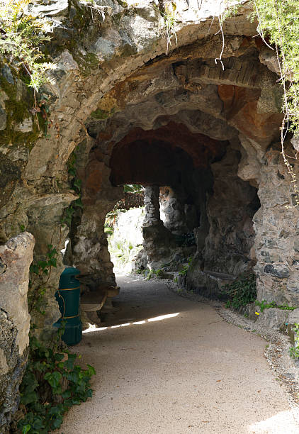 Mysterious stone grotto tunnel in historic Parco di Villa Rocca. Perspective view of artificial rock cavern with arched entrance, stone benches and gravel path leading toward bright sunlight in Mediterranean botanical garden of Chiavari, Italy.
