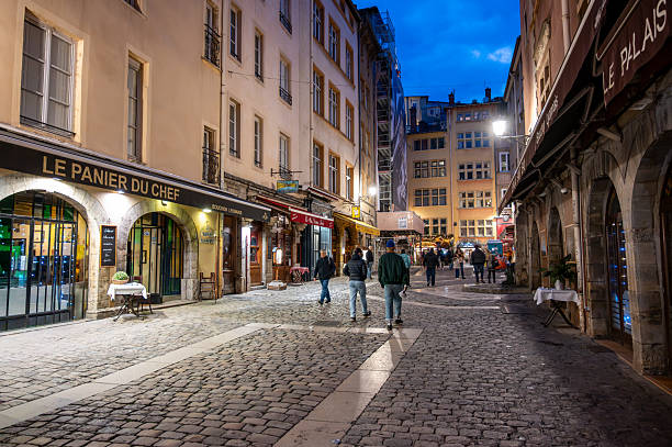 Lyon, france. December 29, 2023. People walking along a cobbled street in historic vieux lyon, france, featuring illuminated buildings and shops at dusk