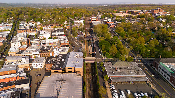 Aerial view of downtown Summit, New Jersey on a spring afternoon. The commuter train station and tracks divide the commercial district with flat-roofed buildings and parking lots from the surrounding residential neighborhoods with mature trees.