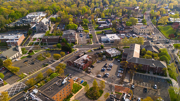Aerial view of downtown district in Summit, New Jersey showing intersection of residential streets with historic church buildings and commercial district. The scene features multiple parking lots, tree-lined streets with spring foliage, and various architectural styles including a prominent stone church. Captured during a spring afternoon.