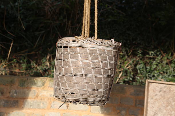 A close-up shot of a traditional, weathered woven bamboo basket suspended in the air by thick, natural fiber ropes. The rustic container hangs against a softly blurred background of dark green garden foliage and a low, earthy stone or brick wall, evoking a sense of traditional rural life and craftsmanship.