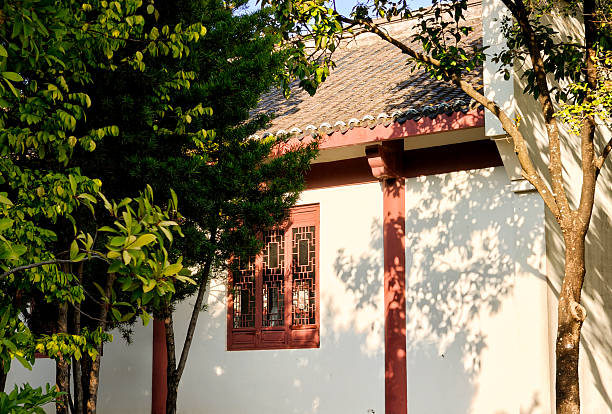 A peaceful, sunlit shot of a traditional Chinese building, where white walls and red wooden details meet a thick green hedge. The bright natural light highlights the rich green tones of the plants, creating a calm, minimalist atmosphere. This image evokes a sense of tranquility and connection to nature, perfect for themes of cultural heritage, garden design, and serene outdoor spaces.
