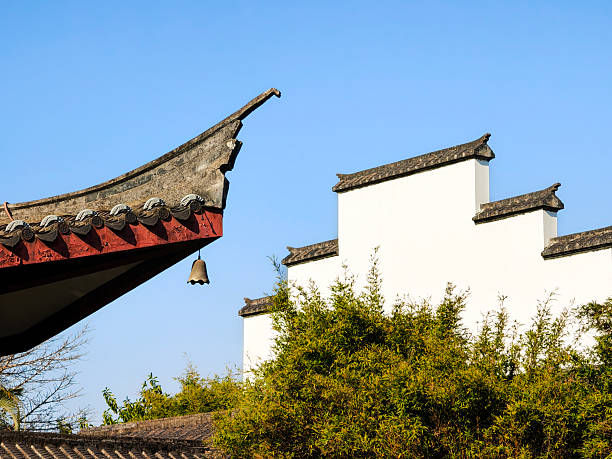 A peaceful, sunlit shot of a classic Chinese roof corner, with its upturned eave and small bell, next to white walls and lush bamboo. The bright blue sky adds a sense of calm and openness, evoking timeless Asian charm. Perfect for themes of travel, heritage, and tranquil garden aesthetics.