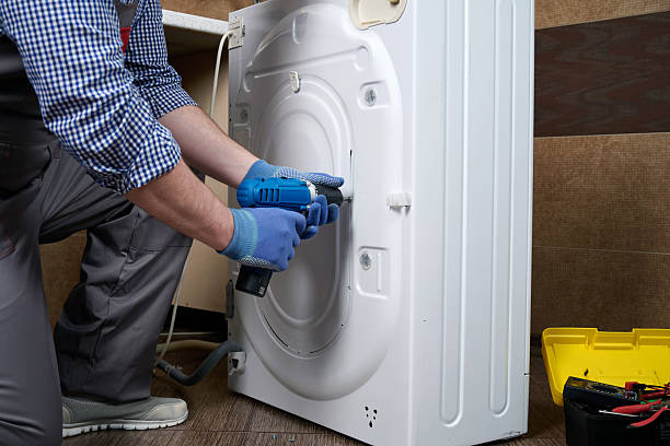 A home appliance repair technician disassembling a washing machine with a power screwdriver.