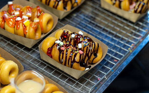Mini donuts topped with chocolate sauce and colorful sprinkles served in a paper tray on a cooling rack. Sweet dessert snack from bakery or street food stall