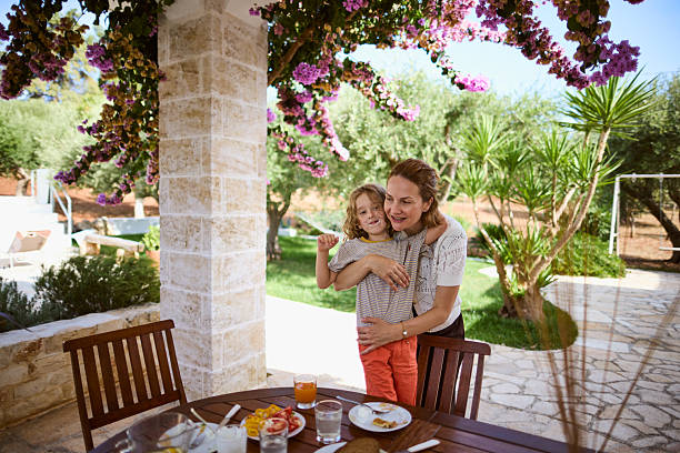 Mother Hugging Child On Sunny Outdoor Patio Breakfast Table In Garden Vacation Home