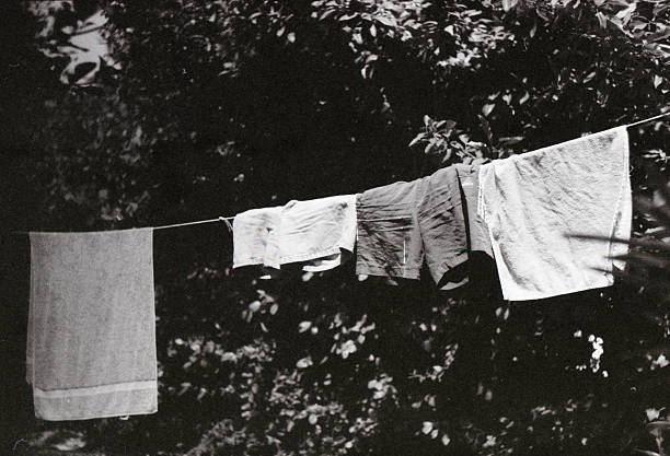 Une photographie argentique authentique en noir et blanc capturant la simplicité d'une scène domestique en extérieur. Du linge de maison, composé de serviettes et de vêtements en coton, est suspendu sur une corde à linge, séchant naturellement devant un arrière-plan de feuillage dense. Le grain naturel du film noir et blanc accentue les textures du textile et le jeu de lumière contrasté entre l'ombre du jardin et l'éclat du linge blanc