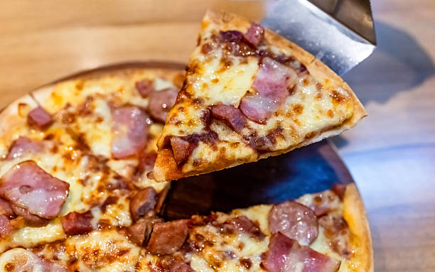 Slice of bacon and cheese pizza being lifted from the tray. Melted cheese and crispy bacon topping creating a delicious fast food and restaurant meal scene