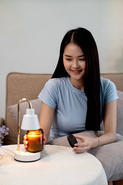 Asian adult woman enjoying cozy evening using electric candle warmer creating relaxing home ambiance self care moment comfortable home living room sofa