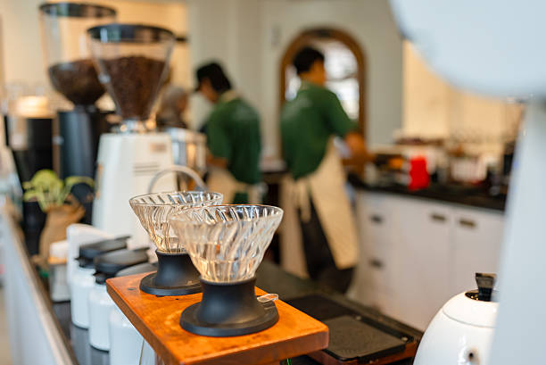 Close-up of empty pour over coffee drippers placed on a wooden stand at the counter in a modern café, with baristas working in the background. The image highlights specialty coffee equipment and the preparation setup in a coffee shop environment.