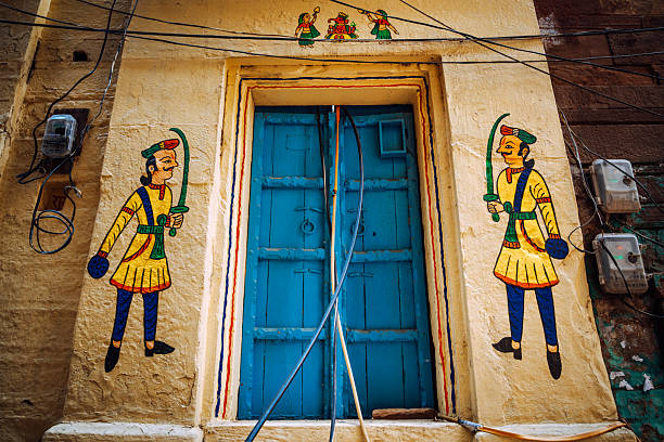 Vibrant mural figures flank a bright blue door on a sunlit wall in Varanasi, India. The scene combines traditional art, architecture, and urban life with bold colors and cultural motifs.