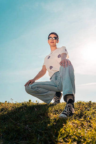Showcasing youthful confidence and self-expression. A young adult woman squats on a grassy hillside wearing sunglasses and casual clothing under a bright sky embodying individuality and a positive outlook.