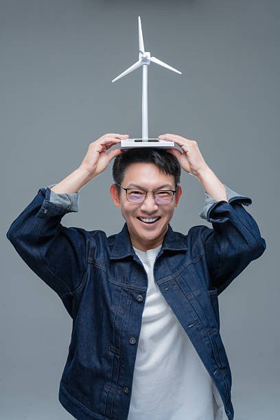 cheerful Asian man with glasses in a denim jacket smiles proudly while holding a model wind turbine with solar panels against a gray background, symbolizing renewable energy, green technology,